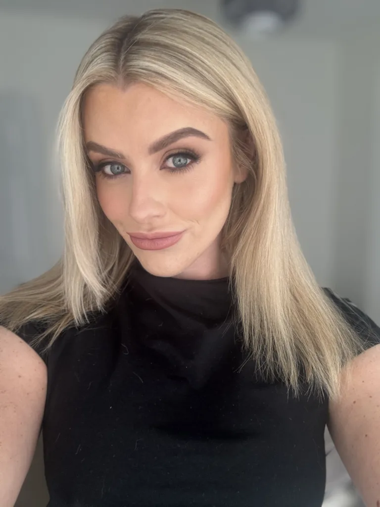Emmacxx poses in a black shiny latex top, showing off her cleavage and blonde hair against a plain white background.
