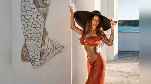 ReyQuins poses in a burnt orange bikini top and matching skirt, leaning against a white wall with a mermaid mural under the sun.