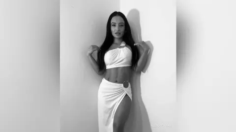 Young woman in a white one-shoulder crop top and matching skirt posing in a minimalist studio setting.