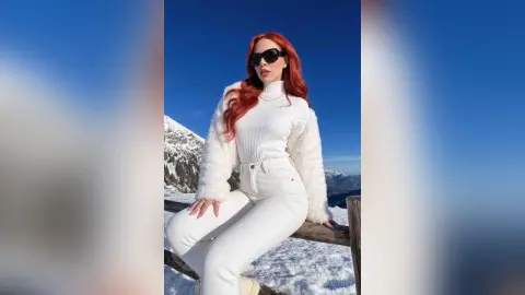 Redheaded woman in a cozy white sweater and pants sitting in a snowy mountain landscape under a blue sky.