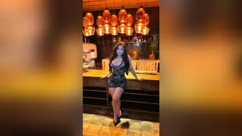 Elegant woman in a black lace cocktail dress posing in a warm, dimly lit upscale lounge with amber pendant lights.