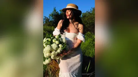 Woman in white summer dress and straw hat holding white hydrangeas in a sunny garden setting.