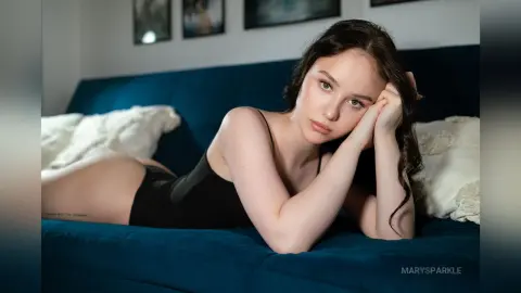 Young woman in a black bodysuit relaxing on a blue velvet sofa in a moody, intimate indoor setting.