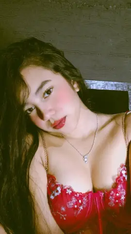 Young woman with long dark hair wearing a red floral lace bralette in a moody, dimly lit indoor setting.