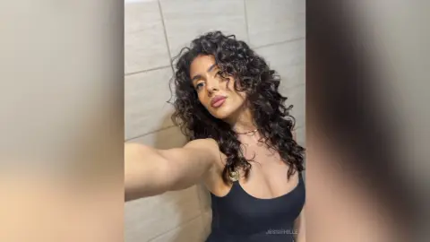 Smiling woman with curly dark hair wearing a black tank top in a modern tiled room.