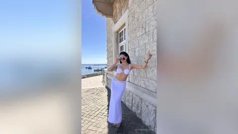 Stylish woman in a white bikini top and long skirt posing by a stone building overlooking the sunny ocean.