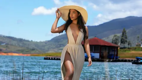 Elegant woman in a white plunging maxi dress and sun hat posing by a scenic mountain lake on a sunny day.