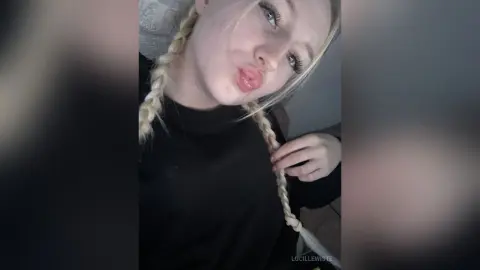 Young woman with blonde braids wearing a black turtleneck making a playful pouty face indoors.