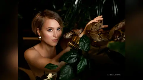 Elegant woman in gold jewelry posing amidst lush tropical leaves in a moody, dark botanical setting.