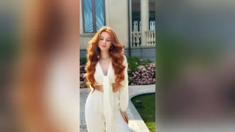 VetaFerr is a redhead in a white crop top and matching pants posing outdoors near a building and flowers.