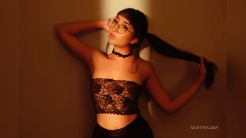 Young woman with glasses and black lace tube top posing in warm moody lighting.