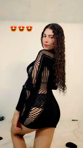 Young woman with long curly hair wearing a black mesh cutout dress in a bright indoor setting.