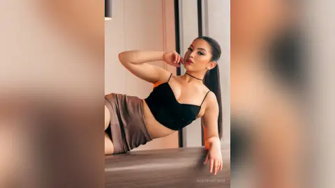 VickyPortman poses in a black bra and brown skirt, leaning back against a surface with her hand near her neck.