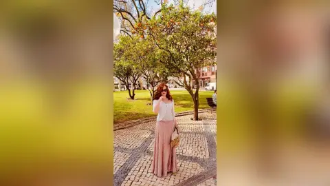Redheaded woman in a white top and long pink skirt standing on a cobblestone path near green trees.