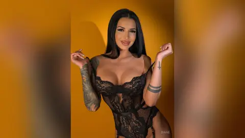 Smiling woman with tattoos wearing black lace lingerie against a warm yellow background.