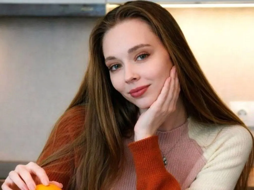 Smiling young woman with long brown hair wearing a pink and orange sweater in a bright kitchen setting.