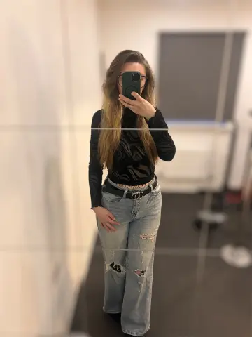 Woman in black turtleneck and ripped light wash jeans taking a mirror selfie in a modern room.