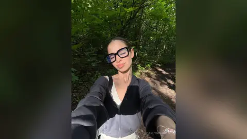 CarlynKernell wearing glasses and a grey zip-up hoodie, posing outdoors in a sunlit green forest.