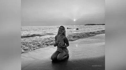 Smiling woman in a patterned sundress kneeling on a beach at sunset in a nostalgic black and white mood.
