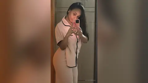 Young woman in cream loungewear taking a mirror selfie in a dimly lit room.