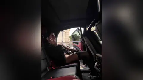 NicoleBella sits in the driver's seat of a car, wearing a black top and black stockings while looking toward the camera.