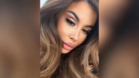 ReynaGomes poses with long, wavy brown hair and heavy eye makeup, looking directly at the camera with a pouty lip.