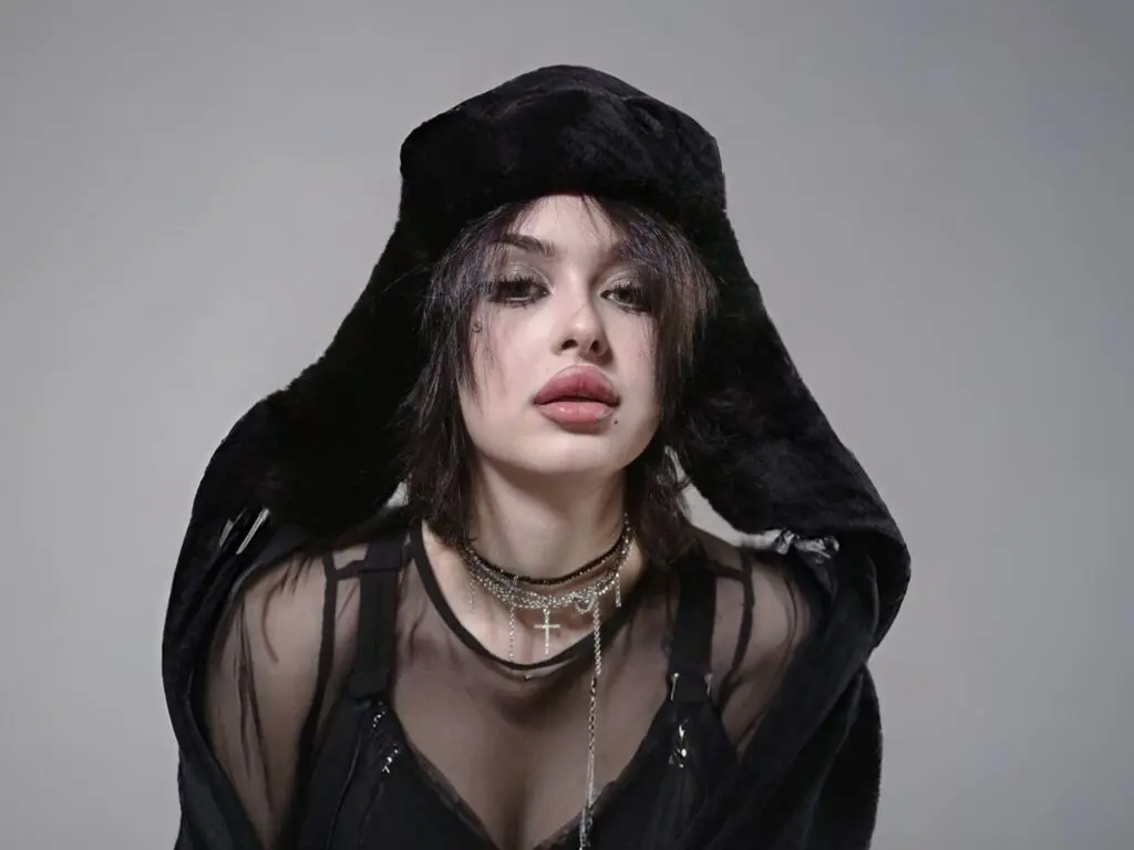 Young woman in edgy black gothic outfit and oversized faux fur hood posing against a grey studio background.