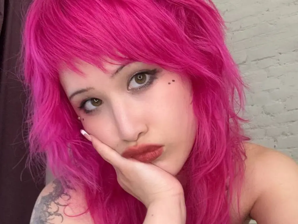 Woman with bright pink shaggy hair and natural makeup posing in a relaxed mood against a white brick wall.