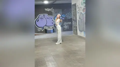 Young woman in white crop top and trousers standing against colorful urban graffiti wall in a moody subway setting.