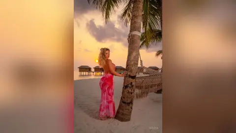 Blonde woman in a pink summer dress leaning against a palm tree on a tropical beach at sunset.
