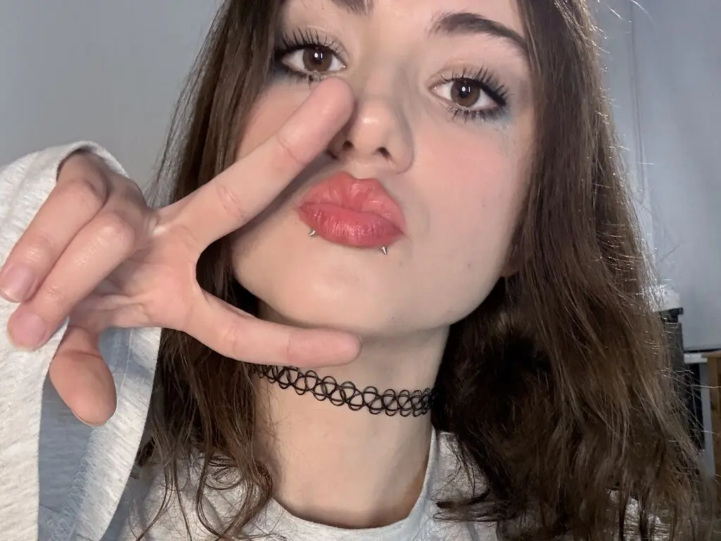 Young woman with lip piercings and a black lace choker making a peace sign in a casual indoor setting.