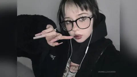 VernellZiccardi wears black glasses and a black hooded jacket, posing with her long pink nails near her mouth while wearing white wired earbuds.