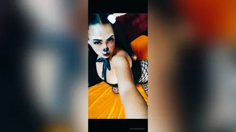Woman in black cat costume with face paint relaxing on a yellow bed in a cozy indoor setting.