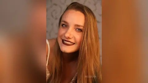 Smiling young woman with long blonde hair and dark red lipstick in a soft, warm indoor setting.