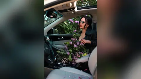 Stylish woman in black leather jacket holding purple lilac bouquet inside a car on a sunny day.