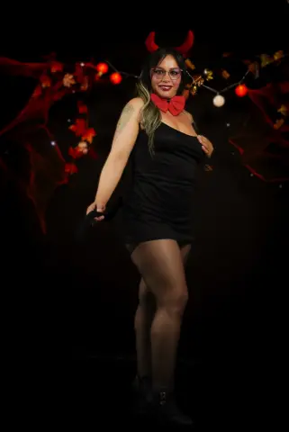 Woman in black dress and red devil horns posing in a spooky Halloween setting with festive lights.