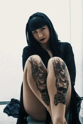 Moody portrait of a woman in a black veil and outfit featuring large skull tattoos on her legs.