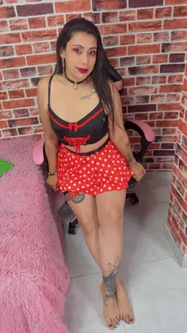 Woman in black lace crop top and red polka dot skirt sitting by a pink fuzzy rug against a brick wall.