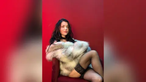Glamorous woman in a white faux fur coat and black lace lingerie posing against a bold red background.