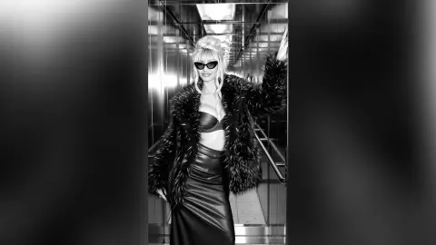 Stylish blonde woman in black sunglasses, leather skirt, and faux fur coat posing in a modern elevator.