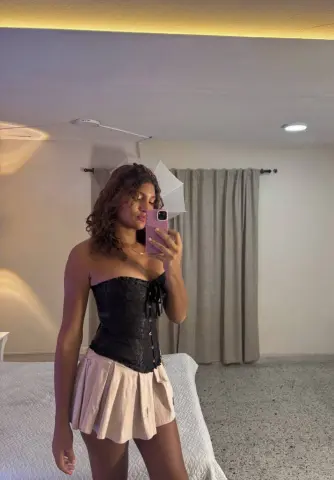 Young woman in a black lace corset and beige mini skirt taking a mirror selfie in a cozy bedroom.