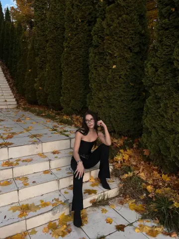 Stylish woman in a black jumpsuit sitting on autumn stairs surrounded by tall evergreen trees.