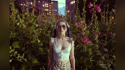 Young woman in a white lace camisole and plaid skirt posing amidst pink hollyhock flowers in a dreamy garden.