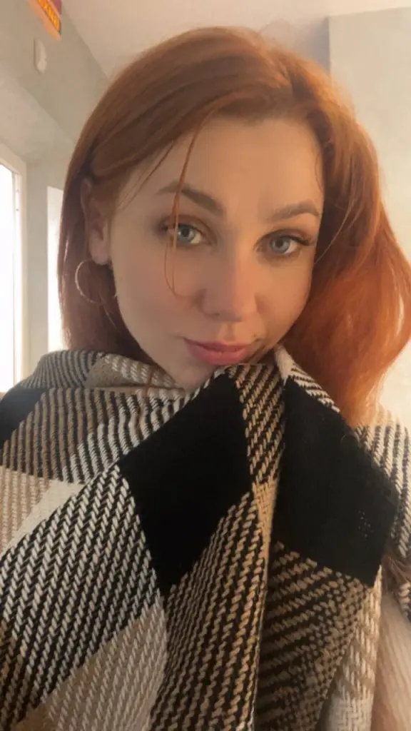 LayaRedfox poses with long red hair, wearing a black long-sleeve top and a pearl necklace against a wooden slatted wall.