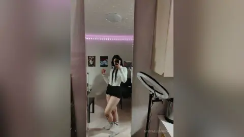 Young woman in a white long-sleeve top and black skirt taking a mirror selfie in a modern bedroom.