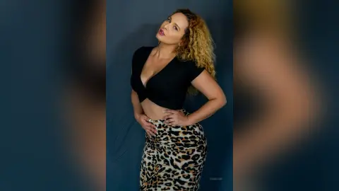 Confident woman in a black crop top and leopard print skirt posing against a blue studio background.