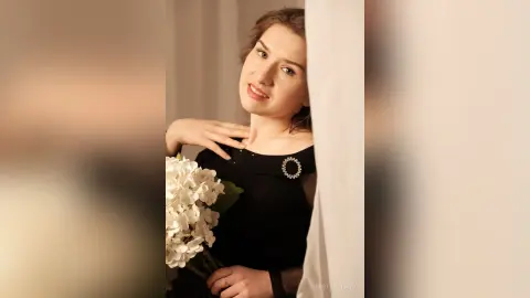 Elegant woman in a black dress holding white flowers by a window in soft, romantic lighting.