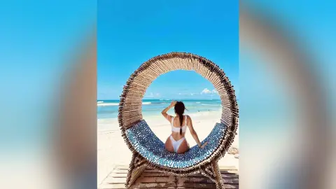 Woman in white bikini sitting in a circular wicker lounge chair on a sunny tropical beach facing the ocean.