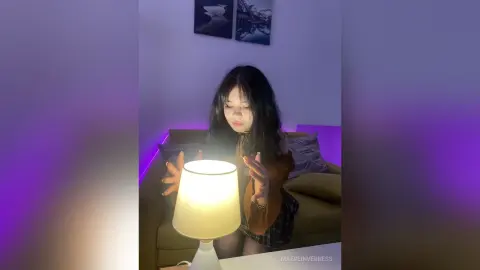 Young woman in a brown top sitting on a sofa by a glowing lamp in a cozy, dimly lit room.