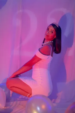 Young woman in a white dress and pearl necklace wearing a tiara, posing in dreamy pink lighting with balloons.
